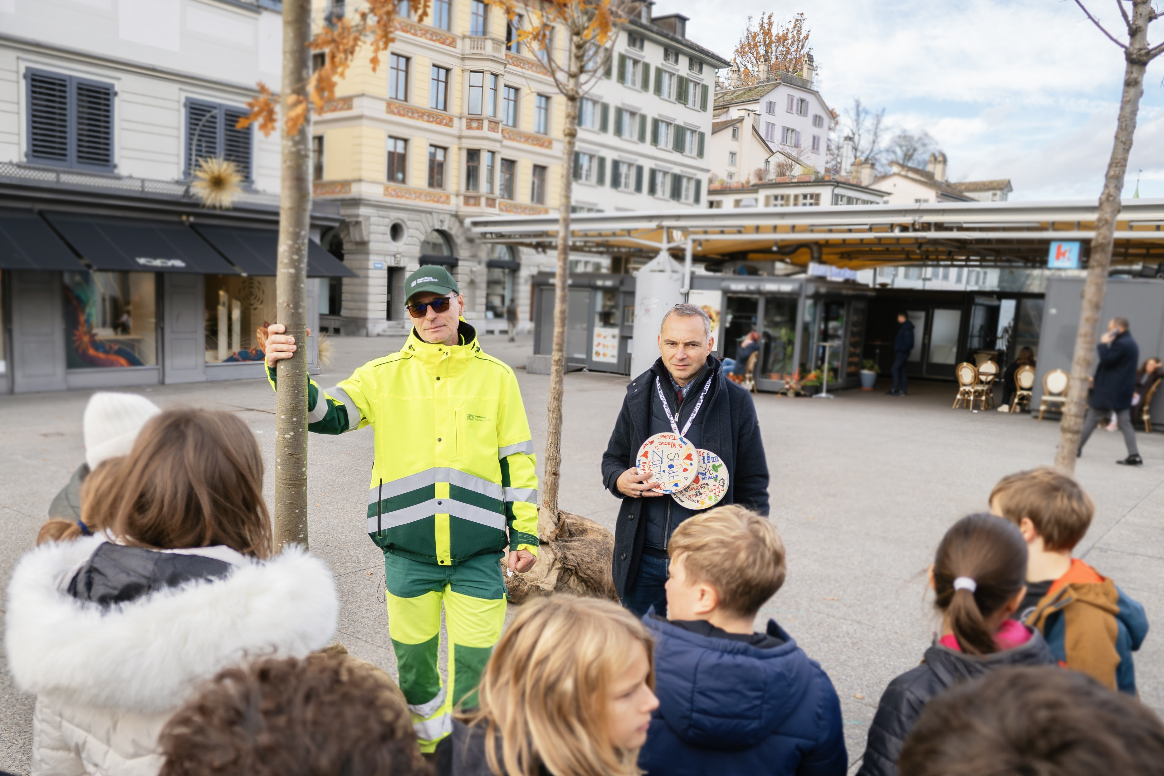 Das Bild zeigt einen Mitarbeiter von Grün Stadt Zürich und einen Mitarbeiter vom KInderdorf Pestalozzi im Gespräch mit einer Gruppe von Kindern.